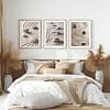 A minimalist bedroom with a bed dressed in beige and cream bedding. Above, three framed Zen Garden Prints featuring abstract stone and sand patterns enhance the serene decor. Pampas grass and soft natural light add to the tranquil ambiance.