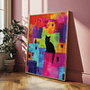 This framed Colorful Cat Art print features a black cat perched on vibrant, abstract rooftops. The artwork rests on a hardwood floor, and sunlight casts soft shadows on a pink wall beside a potted plant.