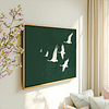 A framed Japandi wall art piece of graceful cranes in flight on a green background is displayed in a cozy room. Sunlight and soft pillows convey a calm, airy atmosphere.