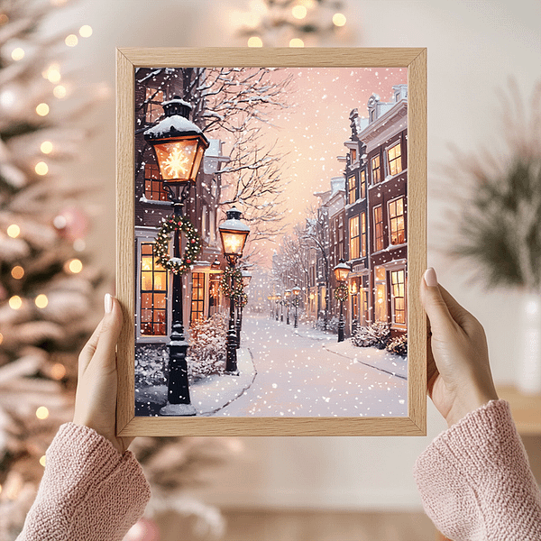 Hands holding a framed painting of a snow-covered street lined with lit lampposts and decorated houses. A Christmas tree with lights is in the blurred background, making this scene feel like a handmade gift from the heart.