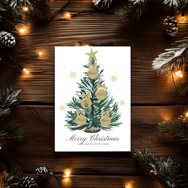 A personalized Christmas card on a dark wooden table features a green tree with gold ornaments showing family names. A pinecone and string lights add to the festive atmosphere.