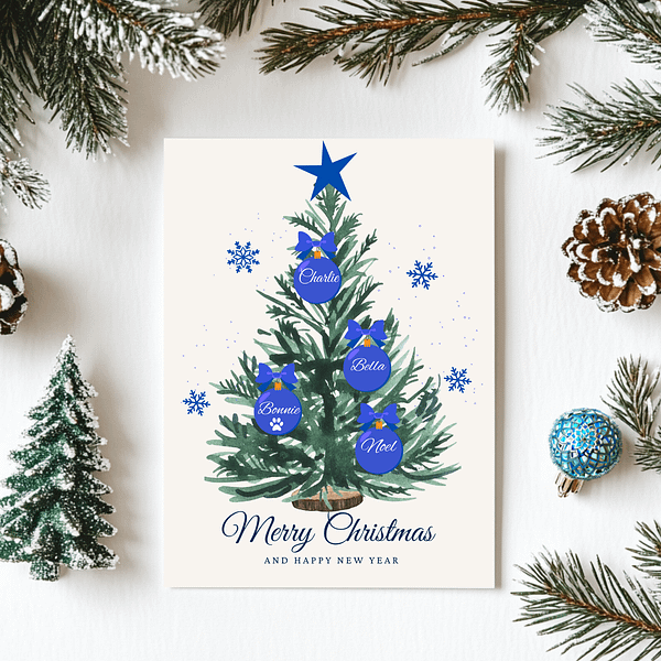 A personalized Christmas card with a green tree and blue ornaments showing family names is set on a white tablecloth with blue-decorated pine branches and a pinecone.